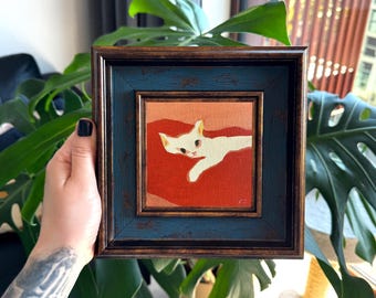 Puede incluir: Pintura enmarcada cuadrada de un gato blanco sobre fondo rojo. La pintura está en un marco de madera azul oscuro y marrón. El gato está acostado con la cabeza girada hacia un lado. El fondo es un degradado de naranja y rojo.