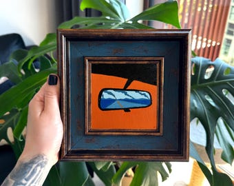 Puede incluir: Una pintura cuadrada en un marco azul oscuro y bronce que representa un espejo retrovisor de coche reflejando una carretera y cielo azules sobre un fondo naranja sólido. La obra presenta una sección superior negra y una vista estilizada.