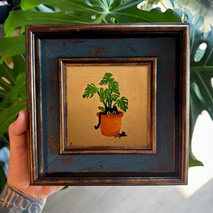 May include: A square painting in a dark blue and bronze distressed frame featuring a black cat with wide yellow eyes peeking out from a terracotta-colored plant pot containing a green monstera plant against a solid gold-toned background.