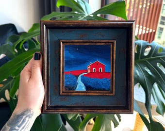 Puede incluir: Una pintura al óleo cuadrada y enmarcada que representa una pequeña casa roja situada en un campo rojo vibrante bajo un cielo nocturno azul oscuro. Un camino sinuoso de color azul claro conduce hacia la casa, que tiene bordes blancos.