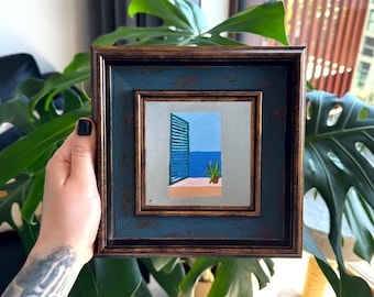 Puede incluir: Un cuadro cuadrado en un marco de color azul oscuro y bronce envejecido. La obra representa una vista a través de una ventana abierta con contraventanas verde azulado, mostrando un mar azul brillante, un suelo de balcón rosa y una planta.