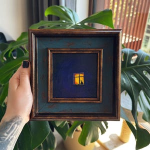 Könnte beinhalten: Ein quadratisches Gemälde in einem dunkelbronzenen und blaugrünen, abgenutzten Rahmen. Das Kunstwerk zeigt eine dunkelblaue Nachtszene mit einem einzelnen leuchtend gelben Fenster, in dem die Silhouette einer Katze auf dem Sims sitzt.