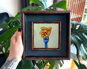 Puede incluir: Una pintura al óleo cuadrada en un marco azul y bronce desgastado que muestra una representación realista de piezas de pasta rigatoni doradas ensartadas en un tenedor de metal azul brillante sobre un fondo verde salvia claro.