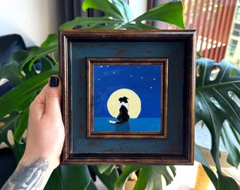 Puede incluir: Un cuadro cuadrado enmarcado que muestra a un perro blanco y negro sentado frente a una gran luna llena amarilla en un cielo nocturno azul oscuro con pequeñas estrellas blancas. El marco es de color marrón oscuro con un acabado texturizado.