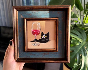 Puede incluir: Obra de arte enmarcada cuadrada que representa un gato negro sosteniendo una copa de vino tinto. El marco combina tonos marrones y azules. Una pieza encantadora para la decoración del hogar.