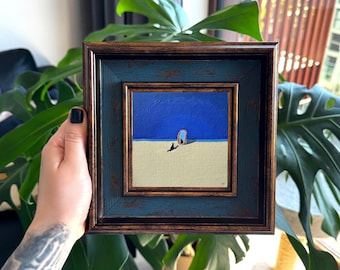 Puede incluir: Una pintura cuadrada en un marco envejecido de color azul y marrón oscuro. La obra muestra a un pequeño gato negro sentado sobre una superficie de arena beige frente a un espejo vertical bajo un cielo azul profundo y sólido.