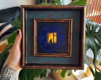 Puede incluir: Pintura enmarcada cuadrada que representa un gato negro silueteado en una ventana iluminada sobre un fondo azul oscuro. El marco es de color verde azulado desgastado con un borde interior marrón. La obra evoca una escena nocturna acogedora.