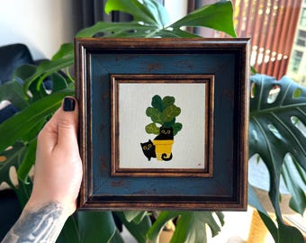 Puede incluir: Una pintura cuadrada en un marco azul oscuro y bronce desgastado que muestra dos gatos negros. Un gato está sentado dentro de una maceta amarilla, mientras que el otro está de pie a su lado. Una planta verde crece de la maceta detrás del gato.