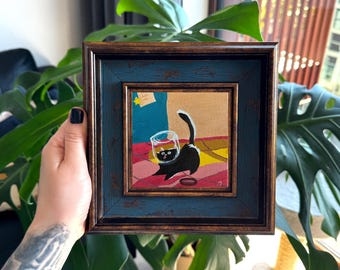Puede incluir: Obra de arte enmarcada cuadrada que presenta una pintura caprichosa de un gato negro con un vaso sobre la cabeza. La pintura tiene un fondo colorido y un marco azul y dorado. La obra de arte se sostiene frente a una gran planta verde.