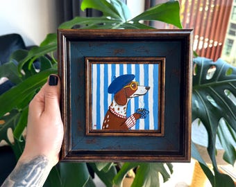 Puede incluir: Obra de arte enmarcada cuadrada que presenta un perro salchicha con boina azul y gafas, sosteniendo una taza de café. La obra de arte tiene un fondo a rayas azules y blancas. El marco es azul desgastado con borde marrón.