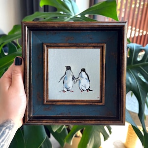 Può includere: Un dipinto quadrato incorniciato su tela raffigurante due pinguini in piedi fianco a fianco su uno sfondo grigio chiaro. I pinguini sono dipinti in nero, bianco e arancione. La cornice è marrone scuro con un bordo interno blu testurizzato.