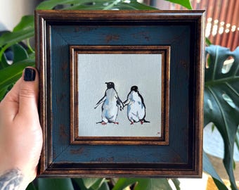 Puede incluir: Un cuadro cuadrado enmarcado sobre lienzo que muestra dos pingüinos de pie uno al lado del otro sobre un fondo gris claro. Los pingüinos están pintados en negro, blanco y naranja. El marco es marrón oscuro con un borde interior azul texturizado.