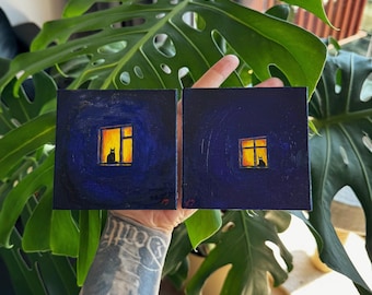 Puede incluir: Dos pequeñas pinturas cuadradas representan la silueta de un gato en una ventana iluminada sobre un fondo azul oscuro. La ventana brilla con tonos amarillos y anaranjados, creando una escena nocturna acogedora. La obra de arte se sostiene frente a una exuberante planta verde.