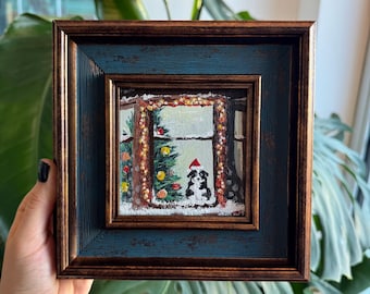 Puede incluir: Pintura enmarcada de un perro con gorro de Papá Noel, mirando por una ventana un árbol de Navidad. La obra de arte está en un marco cuadrado con un acabado azul y marrón, que muestra una escena invernal nevada.