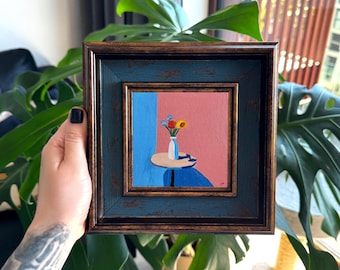 Puede incluir: Un cuadro cuadrado en un marco de color azul oscuro desgastado y bronce. La obra representa un pequeño jarrón blanco con flores coloridas sobre una mesa redonda frente a un fondo dividido en azul y rosa, con una sombra marcada.