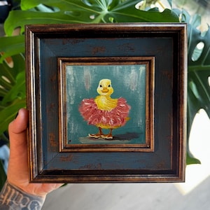 Op de afbeelding: Een klein vierkant olieverfschilderij in een donkere, verweerde lijst met een geel eendenkuiken in een roze tutu met textuur. Het eendenkuiken staat centraal tegen een blauwgroene achtergrond met zichtbare penseelstreken op een donkergrijze ondergrond.