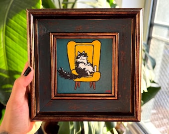 Puede incluir: Obra de arte enmarcada cuadrada que representa un gato blanco y negro descansando en una silla amarilla sobre un fondo turquesa. La obra está en un marco marrón oscuro con un acabado envejecido. El gato tiene una expresión de enfado.