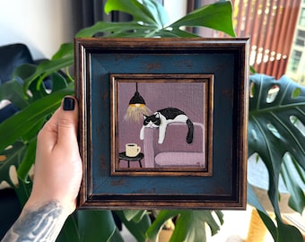 Puede incluir: Pintura enmarcada cuadrada de un gato blanco y negro descansando en un sofá rosa. Una pequeña mesa con una taza de café está al lado del sofá. La pintura está sobre un fondo púrpura con una lámpara colgante.