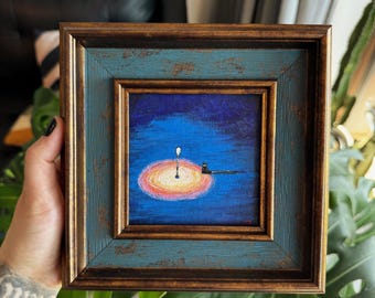 Puede incluir: Pintura enmarcada cuadrada con un marco azul y dorado desgastado. La obra representa un pequeño gato negro sentado sobre una superficie oscura, iluminado por una luz brillante sobre un fondo azul oscuro. La luz está rodeada por un resplandor amarillo y naranja.