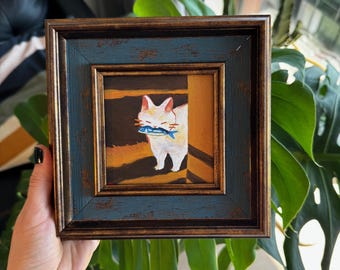 Puede incluir: Pintura enmarcada de un gato blanco con un pez azul en la boca, en una puerta. La obra de arte presenta tonos cálidos de marrón y naranja. El marco es una combinación de azul y marrón.
