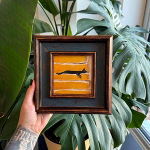 May include: A framed artwork depicting a black cat on a yellow background with white horizontal stripes. The frame is dark blue and brown. The square artwork is held in front of a large green plant.