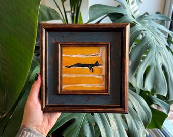 Puede incluir: Una obra de arte enmarcada con un gato negro sobre un fondo amarillo con rayas horizontales blancas. El marco es azul oscuro y marrón. La obra de arte cuadrada se sostiene frente a una exuberante planta verde.