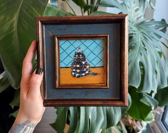 Puede incluir: Pintura enmarcada de un gato negro decorado con luces navideñas y una estrella roja. La obra de arte está ambientada sobre un fondo azul con un patrón de diamantes y un suelo de madera. El marco es una combinación de azul y marrón.