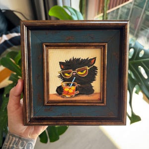 Peut inclure: Tableau encadré d'un chat noir portant des lunettes de soleil et buvant une boisson avec une paille. Le tableau a un cadre bleu foncé et marron. Le chat est représenté avec une boisson dans un verre.