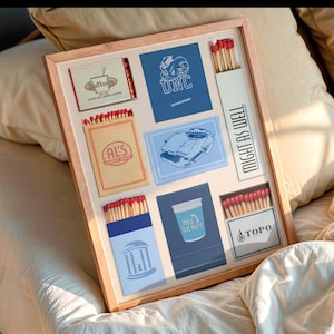 Könnte beinhalten: Ein gerahmtes Kunstwerk mit Streichholzschachteln und Illustrationen. Das Kunstwerk enthält Streichholzschachteln mit rotspitzigen Streichhölzern und verschiedenen Designs, darunter der Text „Might As Well“. Der Rahmen ist hellbraun.