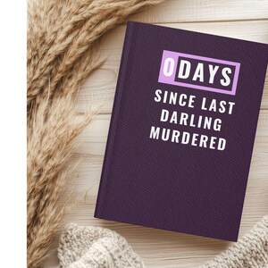 May include: A purple journal with the text "0 DAYS SINCE LAST DARLING MURDERED" in white. The journal is on a wooden surface, next to a cup of coffee and a knitted sweater.