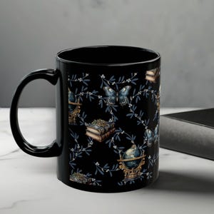 May include: Black ceramic mug with a repeating pattern of butterflies, books, and globes on a black background. The mug has a curved handle and is sitting on a white surface.