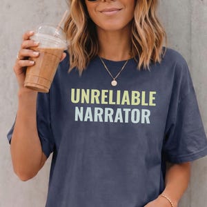 May include: A dark blue short-sleeved t-shirt featuring the text UNRELIABLE NARRATOR printed in two lines. The word UNRELIABLE is in a light lime green font, and the word NARRATOR is in a light blue font, centered on the chest of the shirt.
