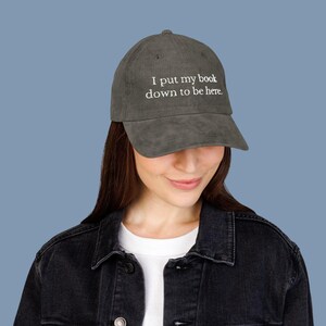 May include: A gray corduroy baseball cap with the embroidered text "I put my book down to be here." The cap is worn by a person wearing a black denim jacket.