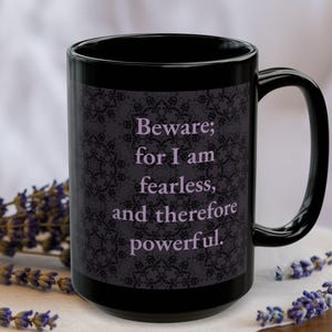 May include: A black ceramic mug with a decorative pattern and the text "Beware; for I am fearless, and therefore powerful." The mug has a large handle and is surrounded by lavender sprigs.