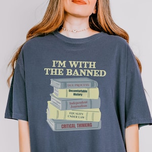 May include: Dark blue t-shirt with the text "I'M WITH THE BANNED" above a stack of books. The books have titles such as "Due Process" and "Critical Thinking". The shirt is a casual style.