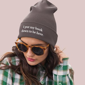 May include: A dark gray beanie with the text "I put my book down to be here." is worn by a person wearing sunglasses and a green and white plaid shirt. The beanie is a knit cap, a type of hat.
