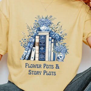 May include: Yellow t-shirt with a graphic featuring a stack of books, blue floral arrangements, and the text "FLOWER POTS & STORY PLOTS" in navy blue. The design has a vintage, watercolor aesthetic.