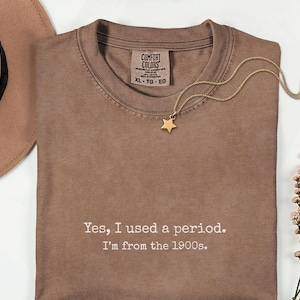 May include: A brown t-shirt with the text "Yes, I used a period. I'm from the 1900s." A gold star pendant necklace and a brown hat are also visible. The shirt has a Comfort Colors tag.