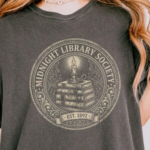 May include: Dark gray t-shirt featuring a circular design with the text "MIDNIGHT LIBRARY SOCIETY" around the edge. The design includes a stack of books, a candle, and the text "EST. 1892" on a banner. The shirt has a vintage, worn look.