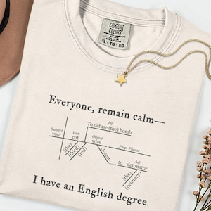 Puede incluir: Camiseta color crema con el texto "Everyone, remain calm - I have an English degree." Un diagrama ilustra la frase. Un collar con colgante de estrella dorada y un sombrero marrón también son visibles.
