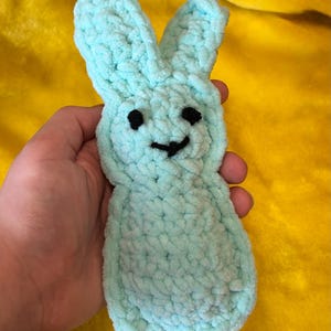 May include: A light blue, plush bunny toy with black eyes and a stitched smile. The bunny is made of a soft, textured material and has long, upright ears. The background is a bright yellow.