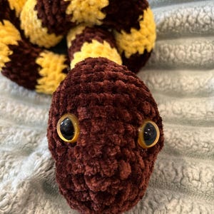 May include: A plush snake toy crafted from soft, fuzzy yarn featuring alternating bands of dark brown and bright yellow. The snake has a rounded head with two large, glossy, amber-colored plastic eyes with black pupils, and a coiled body shape.
