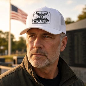 May include: A white trucker cap featuring a rectangular patch on the front. The patch displays a black graphic of an eagle with wings spread above a city skyline and the text NEVER FORGET 9-11-01. The cap has a solid front panel and mesh back.