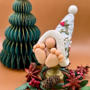 Peut inclure: Une figurine de gnome de Noël fantaisiste avec un chapeau bleu clair et une bordure blanche, posée sur un piédestal doré. Un sapin de Noël en papier vert et des décorations festives entourent le gnome, dont des baies rouges et de la badiane.
