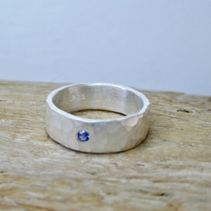 Può includere: Un anello d'argento con una texture martellata e una piccola gemma blu rotonda. L'anello è contrassegnato con il numero "925". L'anello è esposto su una superficie di legno marrone chiaro.