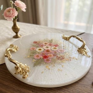 Puede incluir: Una bandeja de servicio redonda de resina blanca con flores prensadas rosas y blancas incrustadas y copos de pan de oro. La bandeja tiene dos asas de metal dorado ornamentadas fijadas en los lados opuestos para su uso.