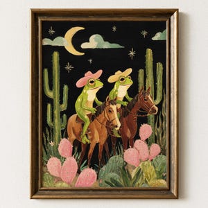 Puede incluir: Impresión artística enmarcada que presenta dos ranas verdes con sombreros de vaquero montando a caballo en una escena desértica. El fondo es negro con cactus, una luna creciente y estrellas. La obra de arte tiene un estilo vintage y caprichoso.