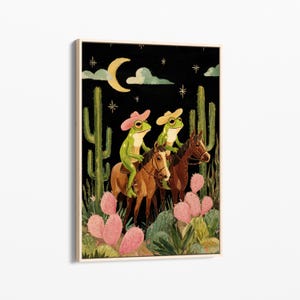 May include: A framed art print depicts two green frogs wearing cowboy hats riding brown horses through a desert landscape. The background is black with a crescent moon, clouds, and stars. Pink cacti and green plants are in the foreground.