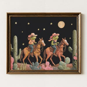 Puede incluir: Una impresión artística enmarcada muestra tres ranas montadas a caballo en un paisaje desértico bajo un cielo nocturno estrellado. Las ranas visten sombreros de vaquero y pañuelos. También se ven cactus y una luna llena.