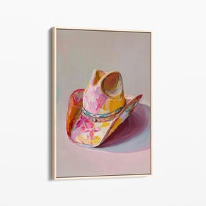 May include: A framed painting of a cowboy hat. The hat is painted with a vibrant mix of colours, including pink, yellow, and orange, with floral accents. The hat's brim casts a shadow on the pink surface below. The artwork is displayed in a light-coloured wooden frame.